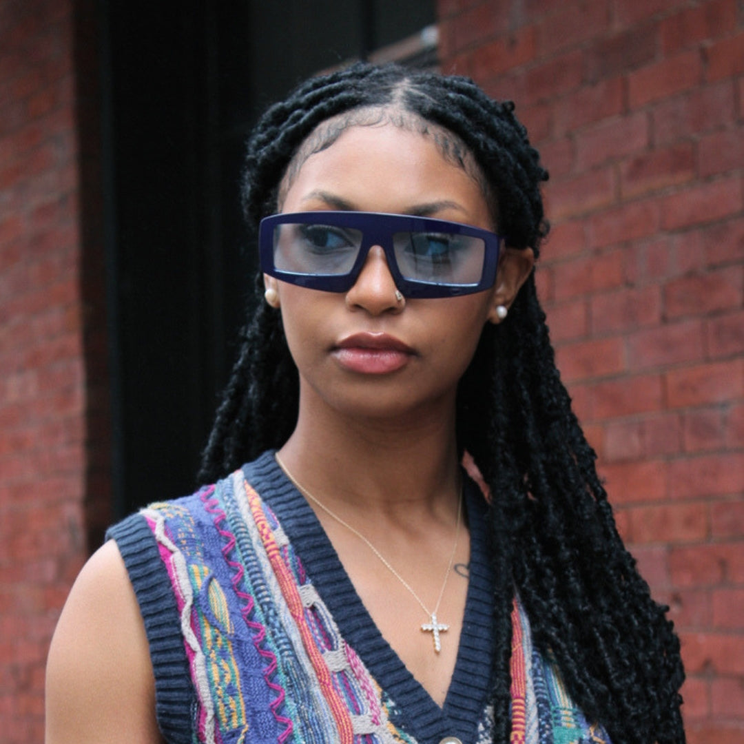 Woman wearing colorful sleeveless top and blue sunglasses in front of a brick wall